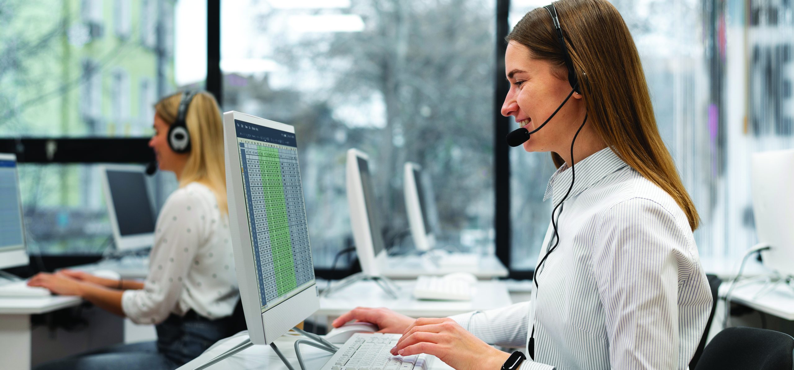 A customer service representative working in a restaurant call center, wearing a headset and typing on a computer, with a notepad and a cup of coffee on the desk.